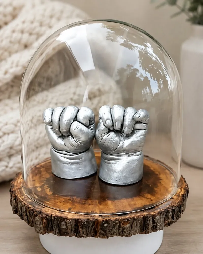 Two tiny baby fists cast in silver, standing on a slice of wood under a glass dome